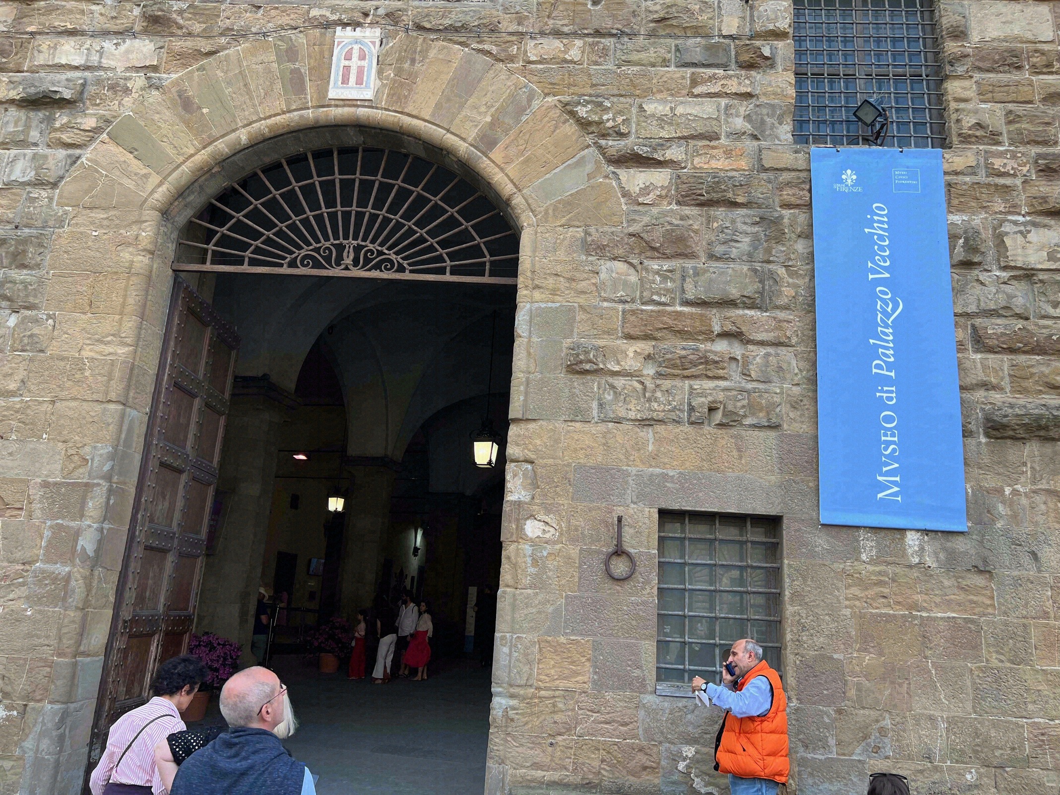 Rustikaler Eingangsportal des Palazzo Vecchio aus Sandstein mit dem blauen Schild zum Museo di Palazzo Vecchio und dem Florentiner Lilienwappen ueber dem Bogen.